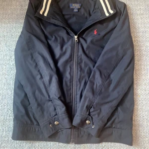 Svart vindjacka Polo Ralph Lauren L - Marinblå vindjacka från Polo Ralph Lauren med dragkedja framtill, klassisk krage med vita ränder och röd broderad logga på bröstet. Jackan har knappdetaljer vid ärmslut och midja. Perfekt för dig som gillar stilren och sportig look. Den passar dig som är runt 157-165 lång. SKRIV OM DU UNDRAR NÅGOT (pris kan diskuteras) ⚠️ONLY SHIPPING TO SWEDEN⚠️