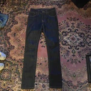 Mörkblå skinny jeans från Nudie - Snygga mörkblå jeans från Nudie Jeans Co med kontrasterande orange sömmar och klassisk femficksdesign. Baktill finns broderade vågmönster på fickorna och en läderpatch med Nudie-logga. Modellen är skinny med smal passform och raka ben. Midja 35 längd 106cm (35)