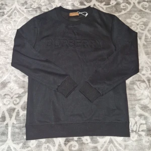 Svart sweatshirt från Burberry - Svart sweatshirt från Burberry med broderad logga framtill. Tröjan har rund hals, ribbade muddar och är tillverkad i mjuk bomull. Perfekt för dig som gillar stilrena och klassiska plagg med exklusiv känsla.