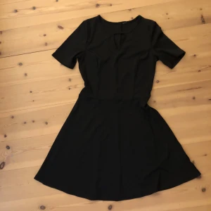 Svart kortklänning med nyckelhålsdetalj - Säljer en svart kortklänning med rund hals och snygg nyckelhålsdetalj framtill. Klänningen har korta ärmar och är tillverkad i ett stretchigt material av 95% polyester och 5% elastan. Enkel och stilren modell som är lätt att matcha.