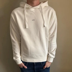 Vit hoodie från A.P.C. med huva - Säljer en stilren vit hoodie från A.P.C. med minimalistisk svart logga på bröstet. Tröjan har huva med breda snören och ribbade muddar vid ärmslut och nederkant. Perfekt för dig som gillar clean och enkel stil. Tröjan är använd 1 gång! Inga defekter eller liknande 