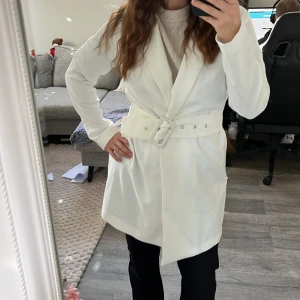 Vit kappa från Gina tricot - Snygg vit kappa med bred krage och matchande bälte i midjan. Jackan har lång ärm och en stilren, rak passform 