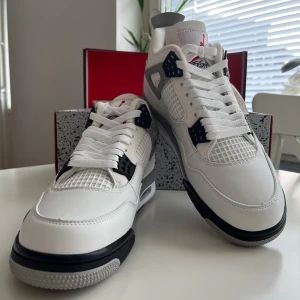 Nike Air Jordan 4 White Cement - Nike Air Jordan 4 White Cement sneakers med vit ovandel i skinn, grå och svarta detaljer samt röd Jumpman-logga på plösen. Snygg meshpanel på sidorna och klassisk snörning. Sulan har gråa stänk och svart kontrast för en riktigt clean look.