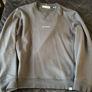 Grå sweatshirt från Les Deux - Säljer en stilren grå sweatshirt från Les Deux med broderad logga på bröstet. Tröjan har rund halsringning, ribbade muddar och är långärmad. Perfekt för en clean och avslappnad look.