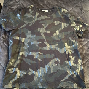 Camouflage t-shirt från Valentino - Snygg t-shirt från Valentino i klassiskt camouflage-mönster med gröna och svarta toner. T-shirten är kortärmad och har rund halsringning. Tillverkad i mjuk bomull för en skön känsla. Perfekt för dig som vill sticka ut med en exklusiv streetstyle-look.