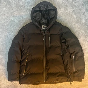 Svart pufferjacka Jack & Jones Core L - Svart pufferjacka från Jack & Jones Core i storlek L. Jackan har huva, dragkedja framtill och flera praktiska fickor med dragkedja. Perfekt för kalla dagar med sin vadderade design och stilrena look. Den är varsamt använt och jag säkjer den eftersom att den inte passar mig längre. Hör av er vid frågor!