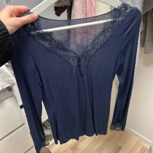 Mörkblå långärmad topp med spets - Snygg mörkblå långärmad topp med v-ringning och spetsdetaljer vid halsen och ärmsluten. Tunn och mjuk i materialet, perfekt för dig som gillar stilrena plagg med en feminin touch. Passar dig som vill ha en enkel men ändå elegant look.