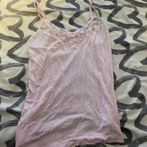Ljusrosa linnetopp med spets - Säljer en ljusrosa linnetopp med tunna axelband och v-ringad hals. Toppen har en snygg spetsdetalj framtill som ger en romantisk vibe. Perfekt för dig som gillar mjuka färger och enkel stil. Materialet känns lätt och skönt mot huden.