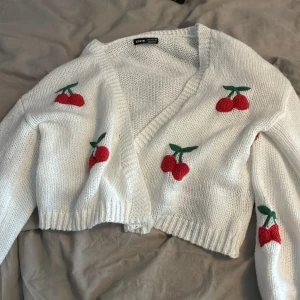 Vit croppad kofta med körsbär från SHEIN - Supergullig vit croppad kofta från SHEIN med broderade körsbär i rött och grönt. Stickad i mjukt material och har långärmad passform med ribbade muddar. Perfekt för att liva upp din outfit med lite färg och lekfull stil.