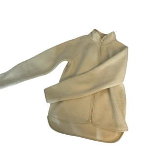 Beige fleecejacka från H&M Move - Mysig beige fleecejacka från H&M Move med hel dragkedja framtill och hög krage. Jackan har lång ärm och är tillverkad i mjuk fleece, perfekt för kyliga dagar. Enkel och stilren design utan mönster eller extra detaljer.