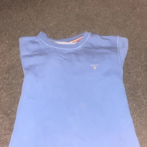 Blå långärmad tröja från Gant - Säljer en stilren blå långärmad tröja från Gant med broderad logga på bröstet. Tröjan är i mjuk bomull och har klassisk rund halsringning med snygg randig detalj på insidan av kragen. Perfekt för dig som gillar enkel och clean stil.