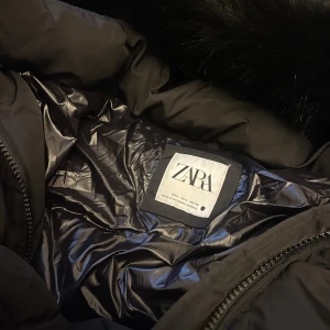 Svart pufferjacka med bälte från Zara - Säljer en svart pufferjacka från Zara med avtagbart bälte i midjan och stor huva med fejkpäls. Jackan har dragkedja framtill och quiltad design som ger en snygg siluett. Perfekt för kalla dagar när du vill vara både varm och stilren. Storlek L men känns som M