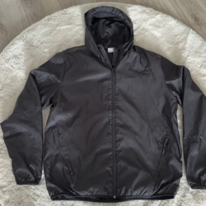 Svart vindjacka med huva från Puma - Svart vindjacka med huva och dragkedja framtill. Jackan har två sidofickor och elastiska muddar vid ärmsluten. Perfekt för blåsiga dagar och enkel att matcha med olika outfits.