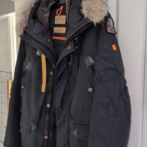 Parajumpers svart parkajacka med päls - Svart parkajacka från Parajumpers, modell Right Hand, med stor huva och lyxig pälsdetalj. Jackan har flera fickor, snygga knappar och dragkedja samt en gul rem vid halsen. Perfekt för kalla dagar och riktigt stilren look.