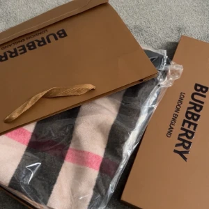 Rutig kashmirhalsduk från Burberry - Exklusiv halsduk från Burberry i 100% kashmir med klassiskt rutigt mönster i beige, svart, grått och rött. Broderad Burberry-logga och ryttare på ena sidan. Mjukt material och fransar i ändarna. Kommer med originalförpackning och taggar.