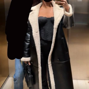 Lång svart pälsjacka med teddyfoder - Säljer en lång svart läderjacka med vitt teddyfoder längs krage, ärmslut och hela insidan. Jackan har en oversized passform och är tillverkad i skinn, fluffigt innerfoder. Perfekt för kalla dagar och ger en riktigt trendig look.