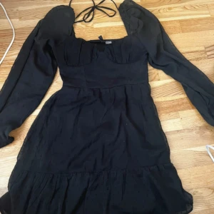 Svart långärmad klänning H&M - Supersöt svart klänning från H&M med långa puffärmar och knytband i nacken. Klänningen har en lätt volang nertill och diskret prickig struktur i tyget. Figurnära upptill och lite utsvängd kjol, storlek i M men ser det som storlek S
