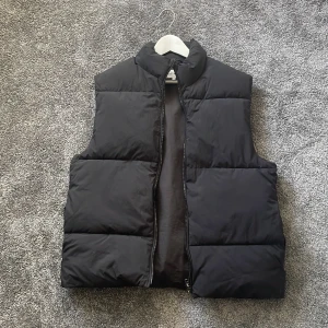 Svart dunväst från East West, M - Svart dunväst från East West i storlek M. Västen har en hög krage och dragkedja framtill. Den är quiltad med breda paneler och har en clean, stilren look. Perfekt att slänga över en hoodie eller tröja när det är kyligt ute. Väster har 1 ficka på varje sida men även ficka inuti som de flesta jackor 