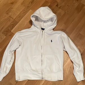 Vit hoodie från Polo Ralph Lauren - Vit hoodie från Polo Ralph Lauren med klassisk blå logga på bröstet. Tröjan har huva med snörning, dragkedja framtill och två stora fickor. Mjuk bomullskänsla och långärmad modell, perfekt för chill dagar.
