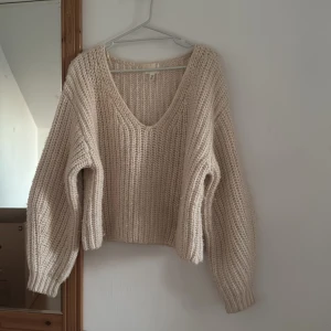 Beige stickad v-ringad tröja H&M - Supermysig beige stickad tröja från H&M med djup v-ringning och breda, långa ärmar. Tröjan har en chunky ribbstickad struktur och en lite croppad passform som ger en chill vibe. Perfekt att slänga på sig när det är kallt ute.