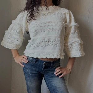 Vit stickad tröja med volangdetaljer - Vit mönsterstickad tröja med trekvartsärmar och volangdetaljer. Tröjan har fransiga ränder och vågig kant nertill och vid halsen som ger en cool look. 