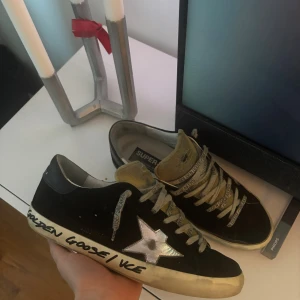 Golden Goose svarta sneakers med stjärna - Svarta sneakers från Golden Goose med vit stjärndetalj på sidan och text på sulan. Skorna har grå snörning med logotyp, beige plös och svart hälparti med märkning. Sulan är platt och har en sliten look som är typisk för märket.