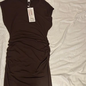Brun omlottklänning med rynk - Säljer en brun klänning med snygg omlottdetalj och rynkade sidor som ger en cool siluett. Klänningen är figurnära med korta ärmar och har en enkel, stilren look. Perfekt för dig som gillar minimalistisk stil och vill ha något som sticker ut.
