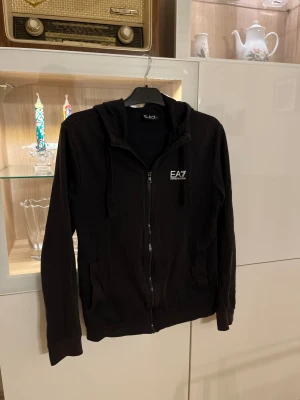 Svart EA7 hoodie med dragkedja - Mörkblå hoodie från EA7 Emporio Armani med vit logga på bröstet. Tröjan har huva med snörning, dragkedja framtill och två fickor. Materialet är mjuk bomull och passformen är normal. Perfekt för dig som gillar stilrena och sportiga plagg.