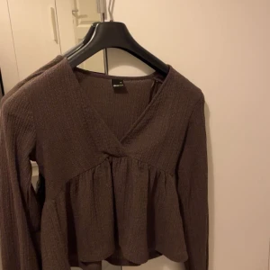 Brun omlott blus från Gina Tricot - Säljer en brun blus från Gina Tricot med omlott-design och v-ringning. Blusen har långärmade ärmar och ett lätt rynkat, flowigt fall nedtill. Materialet är mjukt och har en ribbad struktur som ger en chill vibe.