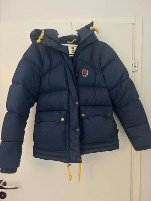 Blå Fjällräven Expedition dunjacka XS - Säljer en blå Fjällräven Expedition Series dunjacka i storlek XS. Jackan har huva, gula snören och klassisk Fjällräven-logga på bröstet. Flera fickor med tryckknappar och dragkedja framtill. Perfekt för kalla dagar och riktigt snygg passform.