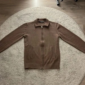 stickad cardigan - Säljer en brun stickad cardigan storlek M. Den har ribbad krage, muddar och fickor samt en hel dragkedja framtill. Perfekt för dig som gillar klassisk stil med lite edge. Materialet är mjukt och skönt, passar till lager-på-lager.