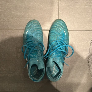 Nike Phantom blå fotbollsskor - Säljer ett par Nike Phantom fotbollsskor i blått med vita detaljer och snörning. Skorna har en stickad ovandel med mönstrad struktur och rund tå. Sulan är utrustad med dobbar för optimalt grepp på planen. Perfekt för dig som vill ha snygga och funktionella skor till fotbollsträningen.