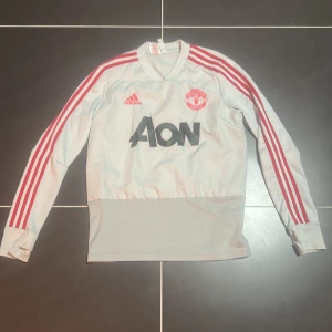 Manchester United långärmad träningströja - Säljer en vit/grå långärmad Manchester United träningströja från Adidas med röda ränder längs ärmarna och klubbmärke på bröstet. AON-logga i svart framtill. Tröjan har en bred mudd nedtill och är tillverkad i ett lätt, syntetiskt material.