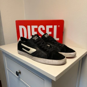 Svarta sneakers från Diesel - Snygga svarta sneakers från Diesel med vita detaljer och röd sula. Skorna har snörning och en stilren design som passar perfekt för en casual look. Diesel-loggan syns tydligt på plösen och sidan av skorna.