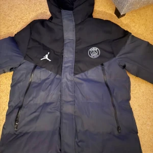 PSG x Jordan lång pufferjacka - Snygg lång pufferjacka från Jordan x PSG i mörkblå och svart. Jackan har huva, två stora dragkedjefickor och loggor på bröstet. Perfekt för kalla dagar med sportig vibe och cool design.