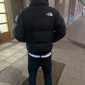 Svart pufferjacka från The North Face - Säljer en svart pufferjacka från The North Face i storlek M. Jackan har en fet, puffig look med hög krage, snörning nertill och dragkedja framtill. Perfekt för kalla dagar och har en clean, urban vibe. Loggan syns tydligt på bröstet och ryggen.