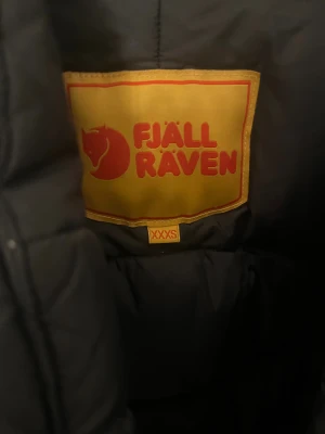 Svart dunjacka Fjällräven XXXS - Svart dunjacka från Fjällräven i storlek XXXS. Jackan har gula snören i huvan, klassisk Fjällräven-logga på bröstet och praktiska fickor med tryckknappar. Puffig modell med dragkedja och knappar framtill. Perfekt för kalla dagar, jackan är en Unisex modell så passar både tjejer och killar, storleken är oxå rätt så stor passar en som har runt xs-s💖finns 2 små slitage som visas på bilderna men inget man tänker på, hör av er om ni har några funderingar 