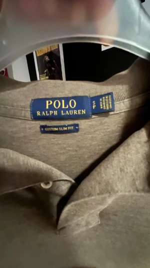 Beige långärmad pikétröja Polo Ralph Lauren - Snygg beige långärmad pikétröja från Polo Ralph Lauren i modellen Custom Slim Fit. Tröjan har klassisk krage, två knappar vid halsen och den ikoniska broderade loggan på bröstet. Perfekt för dig som gillar stilrena och tidlösa plagg.
