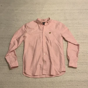Rosa skjorta från Lyle & Scott - Snygg rosa skjorta från Lyle & Scott med klassisk button-down krage och gul broderad logga på bröstet. Skjortan har långa ärmar, bröstficka och knappar framtill. Perfekt för dig som gillar stilrena och fräscha plagg.