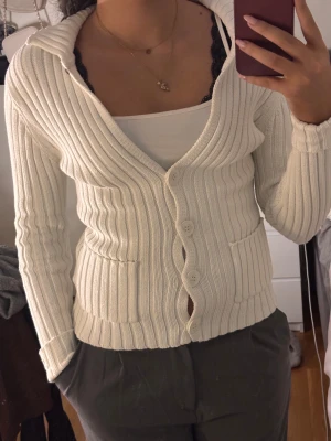 Vit ribbad kofta från GANT - Vit ribbad kofta från GANT med snygg v-ringning och fyra stora knappar framtill. Långärmad modell med två fickor och broderad logga på ärmslutet. Mjuk stickad bomull som sitter normal och har en clean, klassisk look.