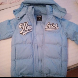 Ljusblå pufferjacka från Hoodrich - Den här jackan är i bra skick jag använde den i vintras och den fungerade mycket bra och den är varm av sig och den är väldig skön och om du vill vara varm i vinter kan du köpa denna jacka jag vill förmedla att den har några fläckar på ärmen o något litet på bröstet men går bort i tvätten. Det ingår ett blått hoodrich snöre med till jackan  