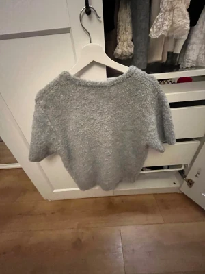 Grå kortärmad kofta från Gina Tricot - Mysig grå kofta från Gina Tricot med korta ärmar och fluffig, stickad känsla. Koftan har knappar framtill och en rund halsringning. Perfekt för lager-på-lager och chill dagar. Snygg att matcha med jeans eller kjol. Stolek M