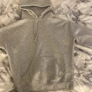 Grå hoodie med magficka och snörning - Säljer en enkel grå hoodie med huva och snörning. Tröjan har en stor magficka framtill och ribbade muddar vid ärmslut och nederkant. Perfekt för chill dagar och passar till allt. Materialet är mjukt och känns skönt mot huden. Dock LITE nopprig men inget man tänker på