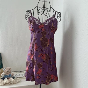 Lila blommig klänning från Victoria’s Secret - Superfin lila kortklänning i satin med blommigt mönster. Klänningen har tunna axelband, spetsdetaljer vid bysten och en lätt utsvängd form. Storlek S, passar bäst 34-36. Dockan har storlek 34.