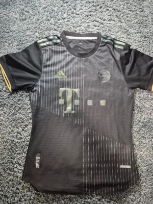 Svart FC Bayern München Adidas tröja - Säljer en svart FC Bayern München fotbollströja från Adidas med korta ärmar. Tröjan har diskreta ljusgula detaljer och ränder på axlarna, samt ett mönster av vita prickar framtill. Materialet är lätt och andas, perfekt för träning eller match.