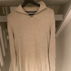 Beige stickad tröja med half zip - Snygg beige stickad tröja med halv dragkedja i halsen. Tröjan har långärmad design och en mjuk, ribbad kant nedtill och vid ärmslut. Perfekt för dig som gillar stilrena och enkla plagg med lite extra detalj vid kragen.