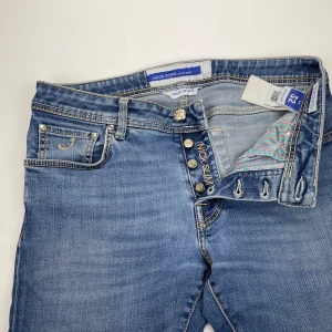 Jacob Cohën jeans  - Snygga blå jeans från Jacob Cohën i modellen Nick Slim. Klassisk femficksdesign med ljusa kontrastsömmar, silverfärgade knappar och en unik etikett i pälsimitation bak. Jeansen har en slim passform och är tillverkade i mjukt denimtyg.