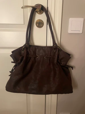 Brun axelväska  med snörning - Snygg brun axelväska i syntetiskt läder med cool snörningsdetalj och metallringar. Väskan har två långa handtag och insidan har flera fack, inklusive ett med dragkedja. Köpt secondhand. Har aldrig använt och säljer den därför! Frakten står på köparen!