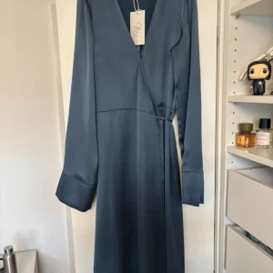 Nakd klänning - Elegant omlottklänning i satin från NA-KD i den vackra färgen Vintage Indigo. Klänningen har långa ärmar med bred manschett, ett knytband i midjan och ett mjukt, glansigt tyg som faller fint. Passar perfekt till fest, middag eller andra finare tillfällen. Kan mötas upp i Stockholm! Nypris 799:-