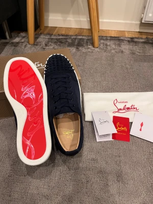 Christian Louboutin mörkblå sneakers - Snygga mörkblå sneakers från Christian Louboutin med ikonisk röd sula och spetsiga nitar på tån. Skorna har snörning, rund tå och är tillverkade i mocka med vita kanter på sulan. Lyxig design med coola detaljer som sticker ut.